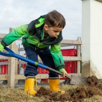 Child in wellingtons digging sand with a large spade